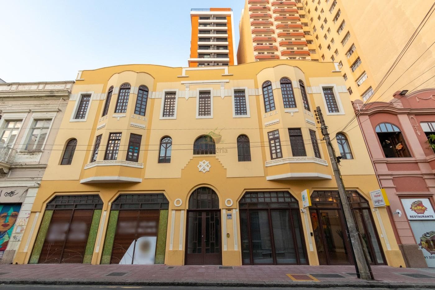 Fachada frontal do edifício em tom amarelado com janelas e portas comerciais no térreo.